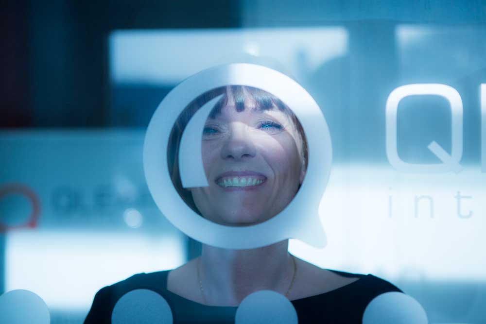 En glad kvinnlig medarbetare på Qlear tittar genom Q:et på logotypen på glasväggen